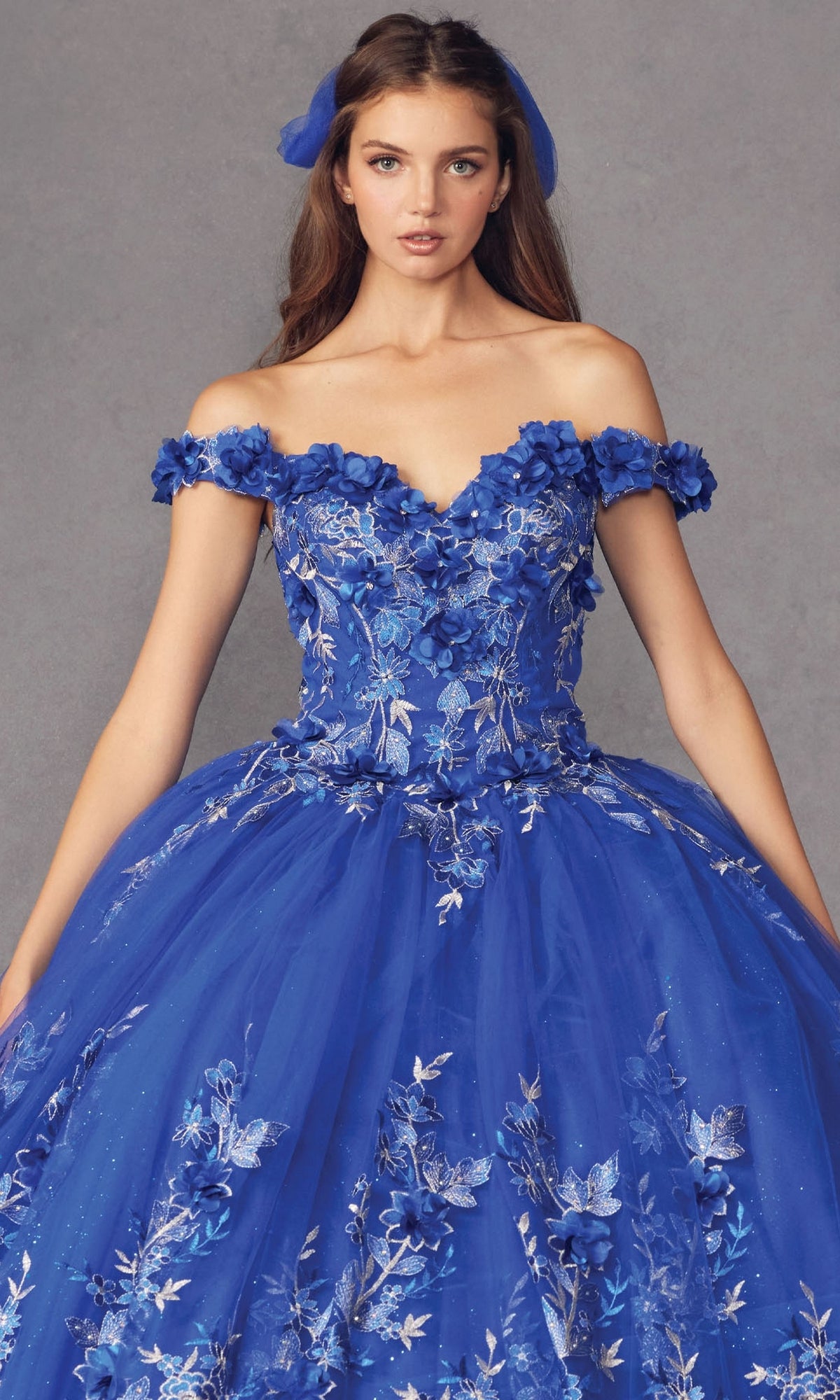 A woman models the Juliet 1447 Off-the-Shoulder Quinceañera Dress in royal blue with beaded embroidery and a matching hair ribbon, posed before a plain gray background.